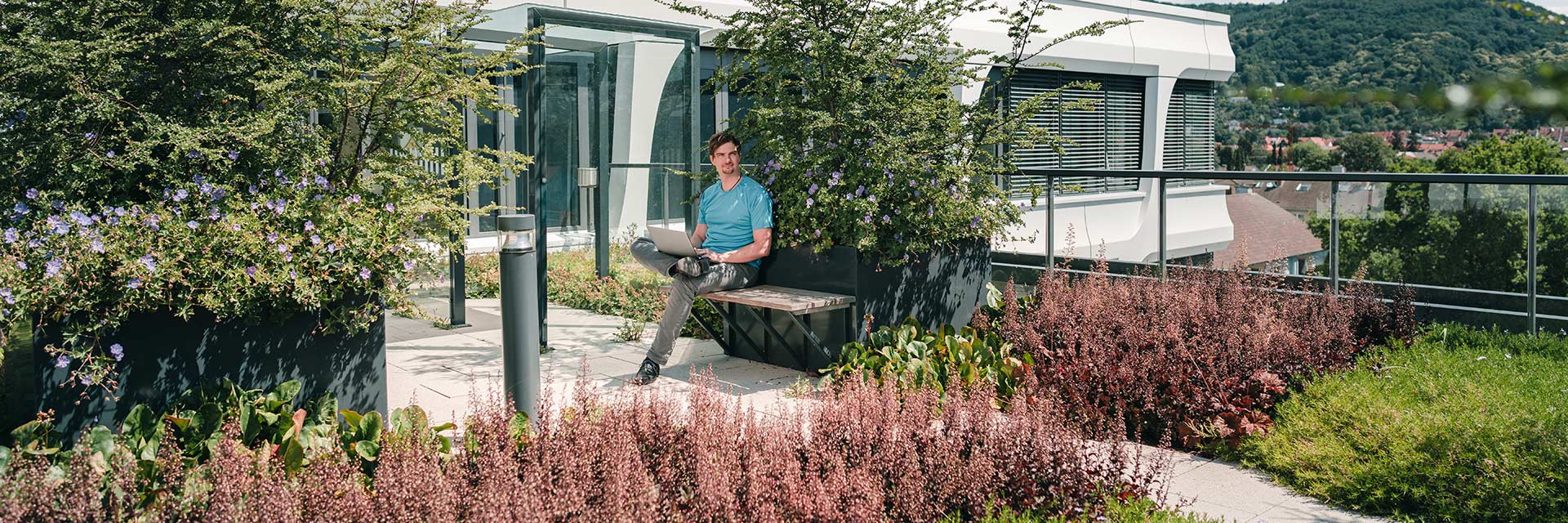 A man with a laptop sits on a green roof terrace, with a white building in the background.