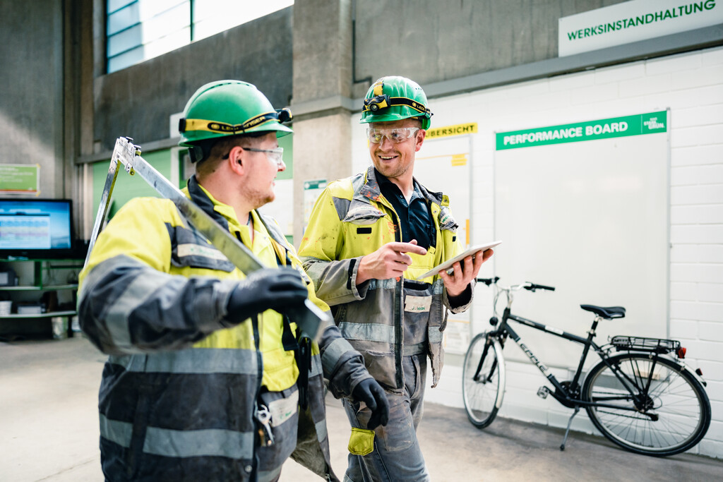 Two men in protective clothing walk through a cement plant, one of them holding a tablet