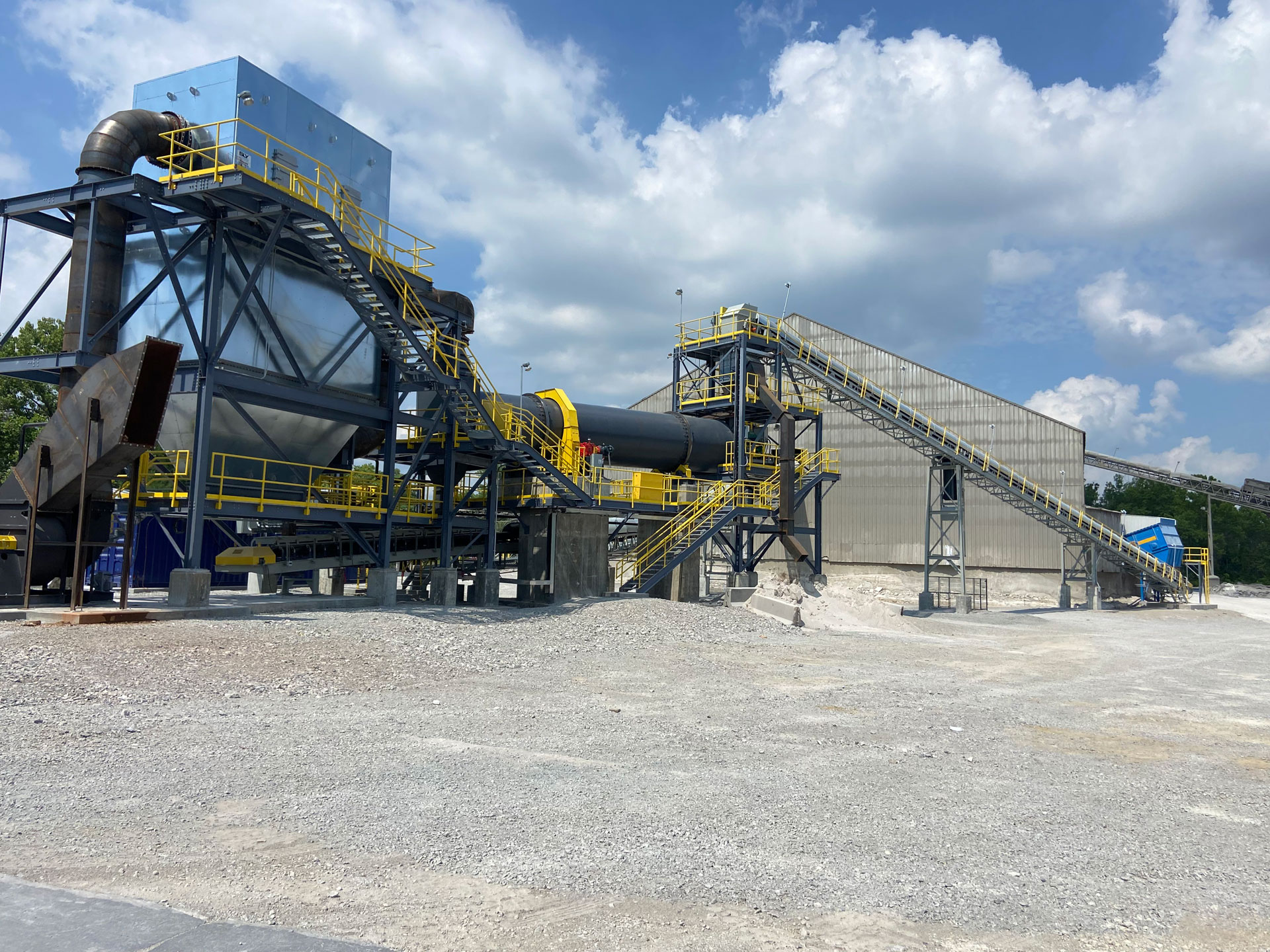 A large slag grinding system in an industrial setting under a cloudy sky. The machinery includes metal platforms, stairs, walkways, and yellow guardrails. A funnel-shaped structure is connected to machines leading to conveyor belts moving ground slag.