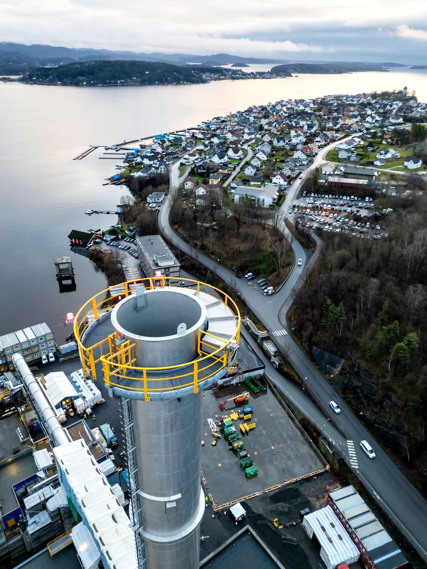 An aerial view of a coastal town with a large industrial chimney in the foreground. The chimney has a yellow safety railing at its top.