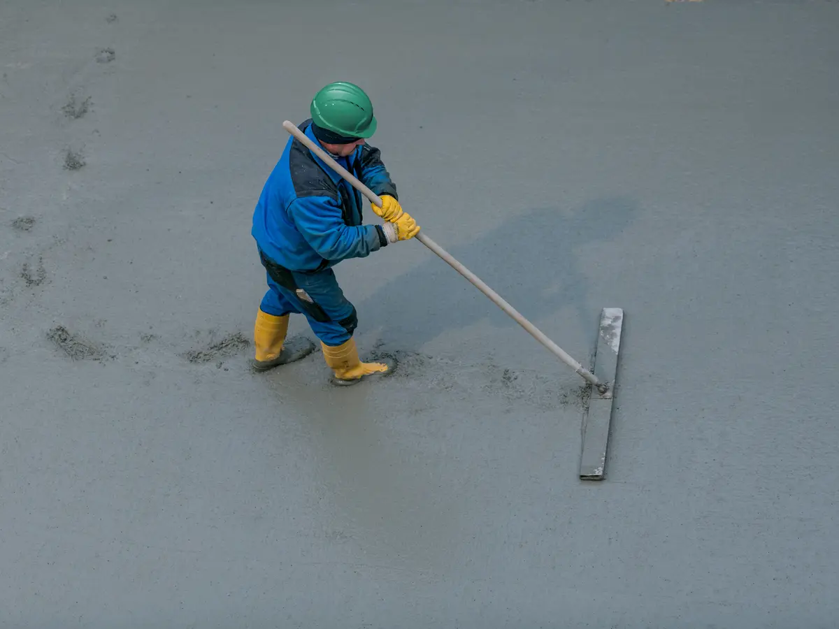 Construction worker in a green helmet, blue jacket, and yellow boots levelling wet concrete with a long trowel on a flat surface.